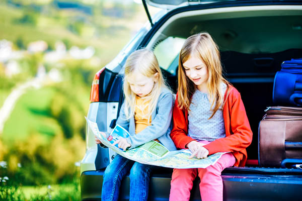 Zwei Kinder sitzen mit einer Landkarte im offenen Kofferraum eines Autos – Lange Autofahrt mit Kindern planen
